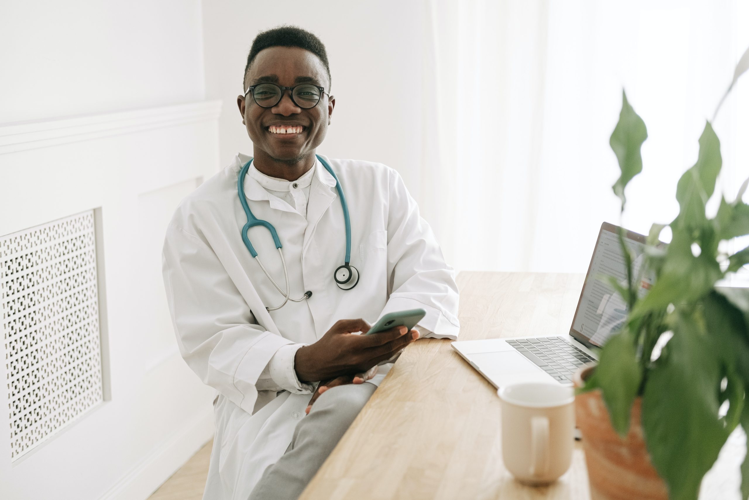 Young male Dr smiling holding his phone setting in front of a laptop with lots of greenery in planters around him.